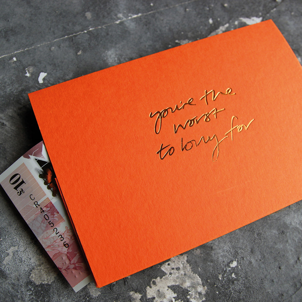 This cash card says You're The Worst To Buy For and is handwritten and hand printed in gold foil on orange luxury paper on grey board