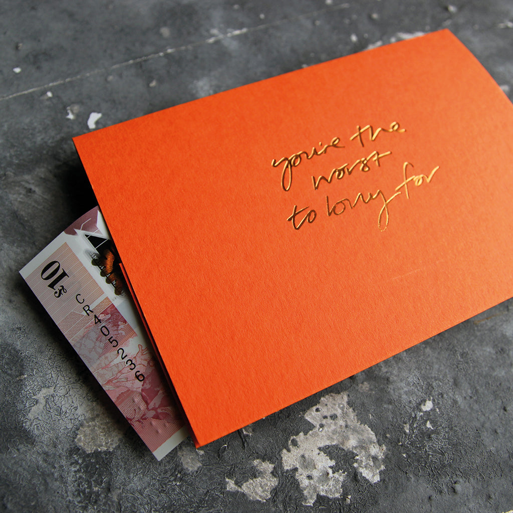 This cash card says You're The Worst To Buy For and is handwritten and hand printed in gold foil on orange luxury paper on grey board