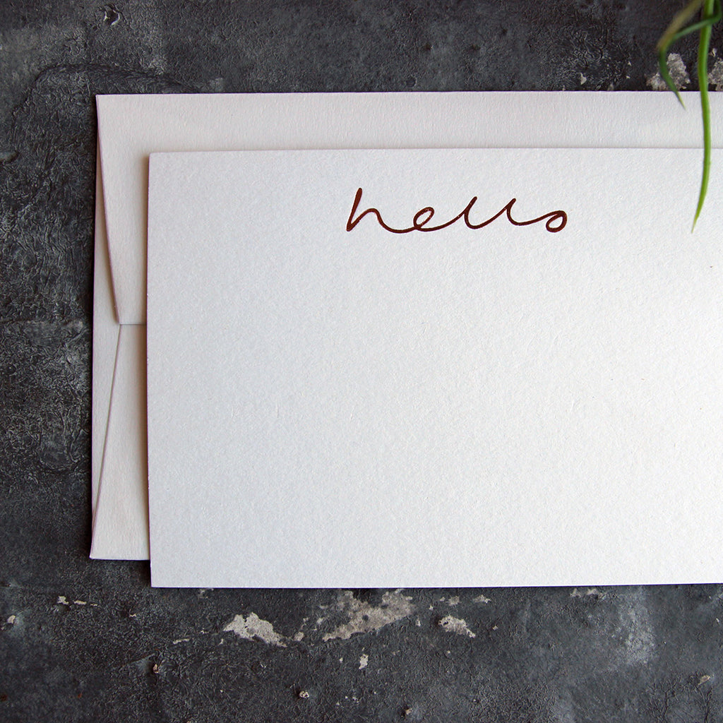 a luxury white notecard which is handfoiled in rose gold foil in handwriting and says hello placed on top of a matching envelope