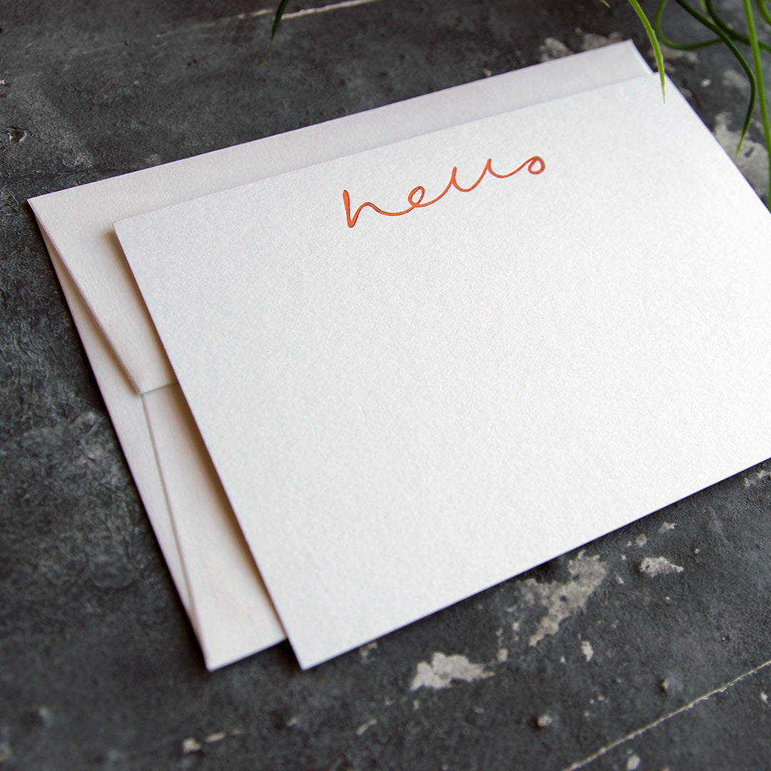 a luxury white notecard and envelope which is handfoiled in rose gold foil in handwriting and says hello placed on top of a matching envelope