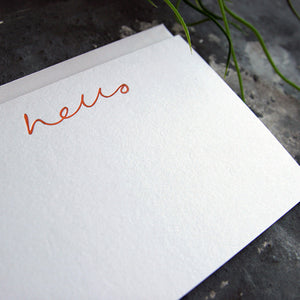a close up of a luxury white notecard which is handfoiled in rose gold foil in handwriting and says hello placed on top of a matching envelope