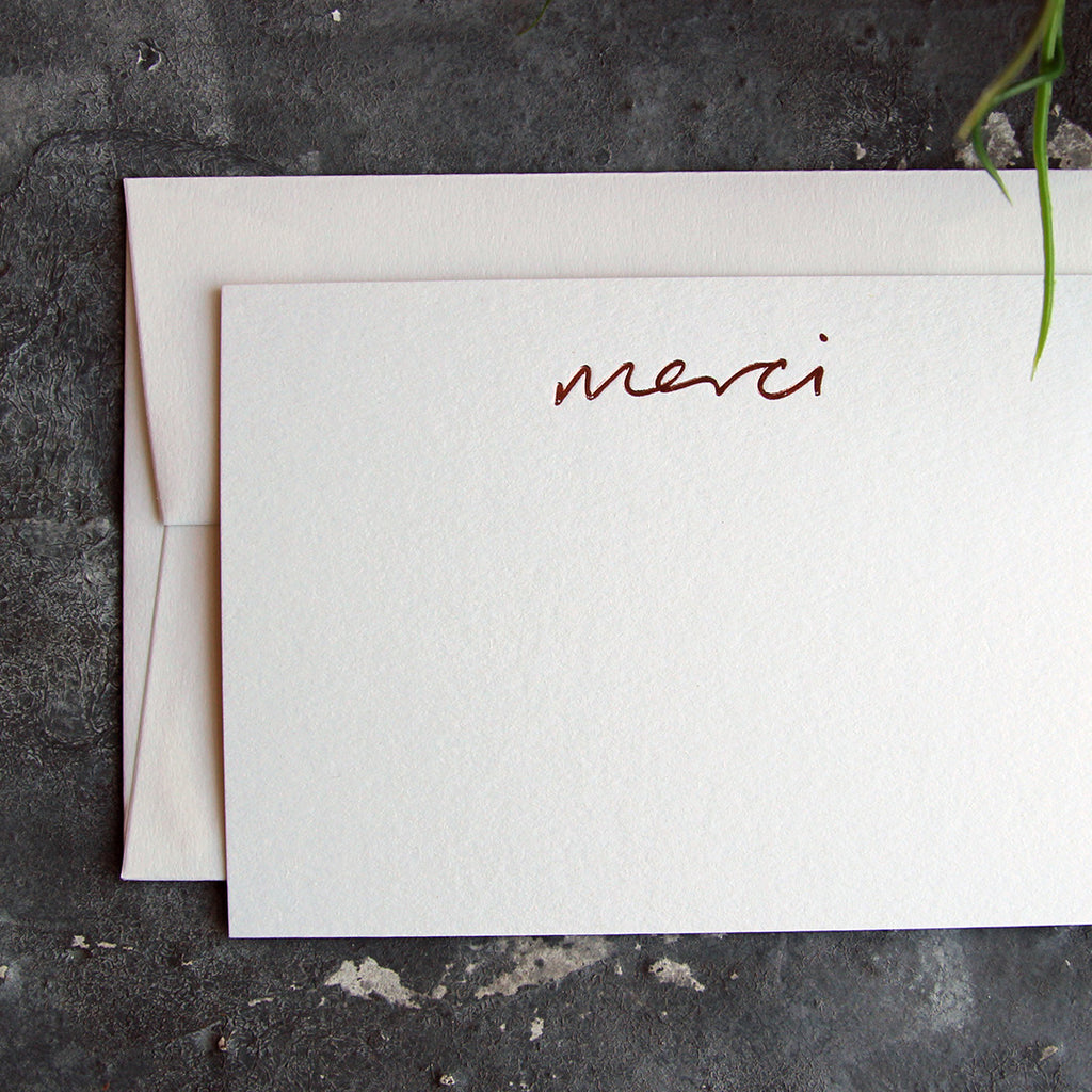 a luxury white notecard which is handfoiled in rose gold foil in handwriting and says merci placed on top of a matching envelope