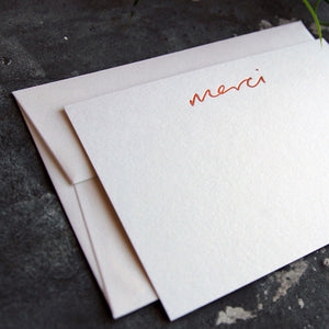 a luxury white notecard which is handfoiled in rose gold foil in handwriting and says merci placed on top of a matching envelope
