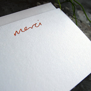 a close up of a luxury white notecard which is handfoiled in rose gold foil in handwriting and says merci placed on top of a matching envelope