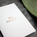 a luxury handwritten rose gold hand foiled message says Oh Merde on the front of the card