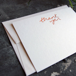 a close up of a luxury white notecard which is handfoiled in rose gold foil in handwriting and says thank you placed on top of a matching envelope